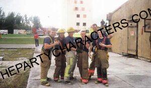 Group of seven firefighters in turnout gear posing together outdoors at a training site, some with helmets on.